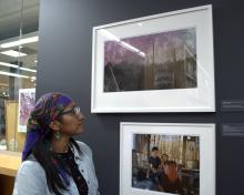 Amber Lee Ortega wears a purple headscarf with a multicolored, floral print, as well as glasses, turquoise earrings, a black printed shirt, and a light blue button-down. She looks at her photographs framed on a dark gray wall, one of which features a desert landscape with saguaro cactus, and another that centers three people in a house. 