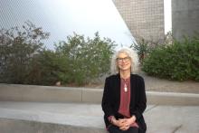 Brenda Hillman sits on a low gray wall in front of a sidewalk area, behind which there are various desert plants and a slanted steel wall. She wears glasses, a maroon shirt, black cardigan, and various silver necklaces.