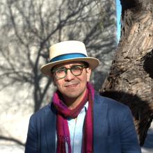 Edgar Garcia stands in front of a tree and wears a hat with a blue ribbon around the base, glasses with green frames, a navy blue blazer, a light blue button-down shirt, and a maroon scarf.