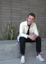 Edgar Kunz sits on a gray bench wearing black jeans, purple socks, white sneakers, and a white unbuttoned button-down. He sits in front of a gray brick background next to a plant.