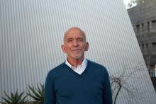 Mark Doty wears a blue sweater over a white shirt and stands in front of a white wall with plants visible behind him.