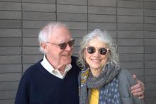 Robert Hass has his arm around Brenda Hillman as they stand smiling in front of a gray brick wall. Robert Hass is to the left and wears a navy blue sweater with a collared shirt underneath. Brenda, to the right, wears a yellow shirt, patterned scarf, and gray cardigan. Both wear sunglasses.