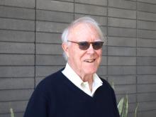 Robert Hass stands in front of a grey brick wall while wearing sunglasses, a white collared shirt, and a navy blue sweater.