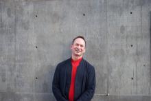 Forrest Gander wears a red shirt beneath a navy blue jacket and stands in front of a grey wall smiling.