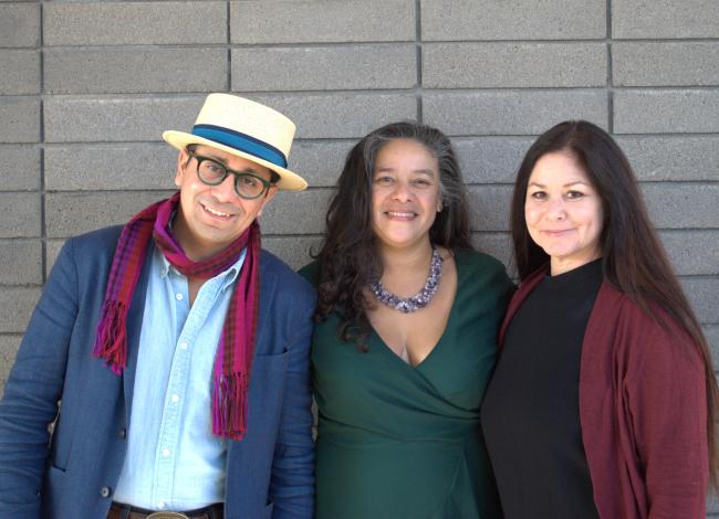 Edgar Garcia, Sheila Maldonado, and Gina Franco stand in a row in front of a grey background. Edgar Garcia wears a hat with a blue ribbon around the base, glasses with green frames, a navy blue blazer, a light blue button-down shirt, and a maroon scarf. Sheila Maldonado wears a dark green wrap dress and a necklace of purple crystals. Gina wears a black dress and maroon cardigan.