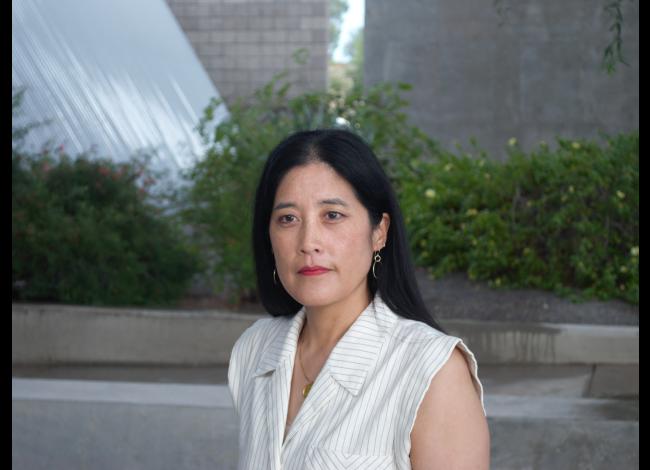 Victoria Chang wears a white shirt and poses for a photograph in front of a garden with grey walls in the background.