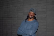 Jericho Brown wearing a grey-blue sweater smiles with his arms crossed