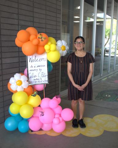 Celia Pérez wears a brown dress with black vertical stripes, black sneakers, black-framed glasses, and a gold necklace. She stands to the right of a pink, yellow, blue, and orange balloon display with balloon daisies and a sign that reads, "Welcome to a matinee reading by Celia C. Pérez author of "The First Rule of Punk."" Both Celia and the balloon display are in front of a grey brick and glass backdrop.