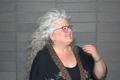 Dana Levin wears a black robe with slight yellow, blue, green, and orange embroidery over a black undershirt, and silver bracelets. She wears black and purple-rimmed glasses and gold loop earrings. She is looking towards the right side, and the wind sweeps her curly hair back from her face. She stands in front of a grey brick wall.