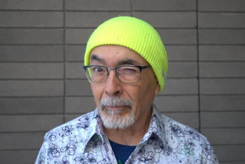 Juan Felipe Herrera winks in front of a gray brick wall. He wears a highlighter yellow beanie, glasses, and a printed button-down t shirt.