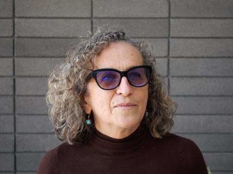 Liliana Valenzuela poses in front of a gray brick wall wearing a brown turtleneck, sunglasses, and blue gem earrings.