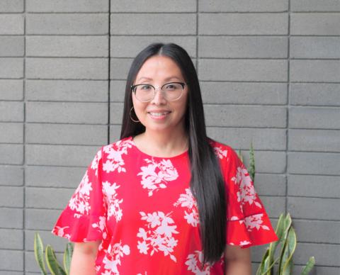 Mai Der Vang poses in a red and white floral top in front of a grey wall. Some green plants peek out in the background, and Vang, wearing glasses, is smiling.