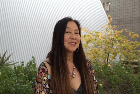 Marilyn Chin wears a necklace with large silver charms and a multicolored cardigan. She stands before an angled steel wall bordered by green plants, one of which has yellow flowers blooming.