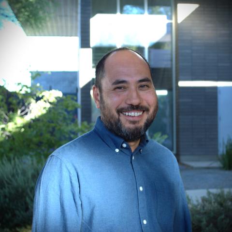 Brandon Som stands wearing a blue button down shirt in front of a grey brick building with long glass windows.