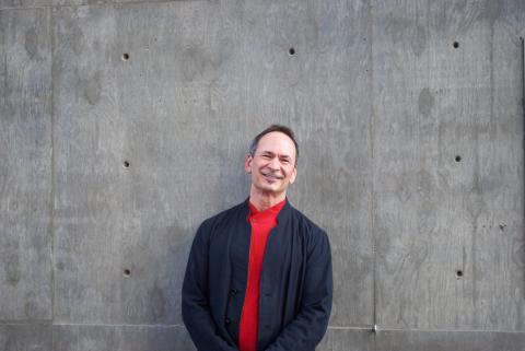 Forrest Gander wears a red shirt beneath a navy blue jacket and stands in front of a grey wall smiling.