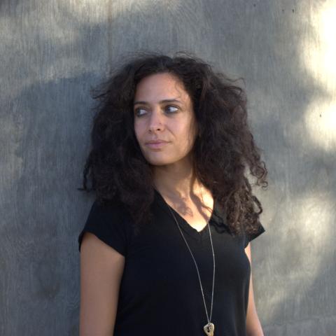 Hala Alyan wears a black v-neck t-shirt and a long gold necklace. She stands in front of a grey wall.