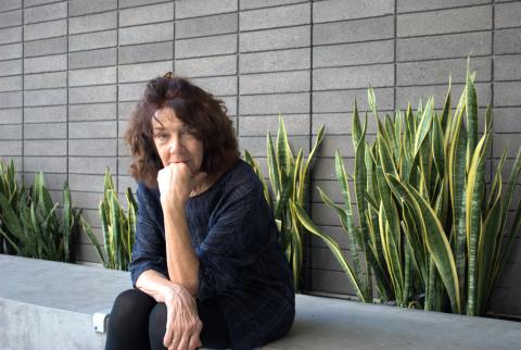 Mary Ruefle wears a blue plaid blouse and black tights. She sits cross-legged on a gray stone bench and her chin rests on the back of her knuckles as she faces the camera. She sits before a grey brick background with a row of plants behind her.