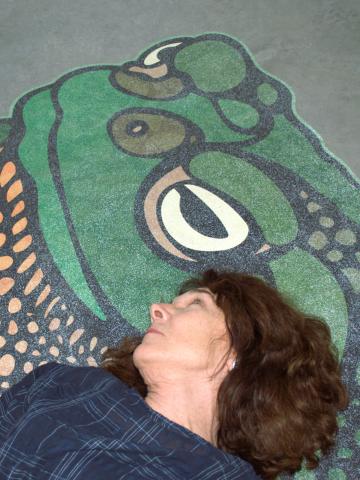  Mary Ruefle lays down on the floor on top of a large green sticker art piece of a sonoran desert toad. Mary's looks up, as if looking in the eyes of the toad.