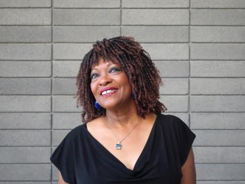 Rita Dove smiling and wearing a black shirt in front of a grey brick wall.