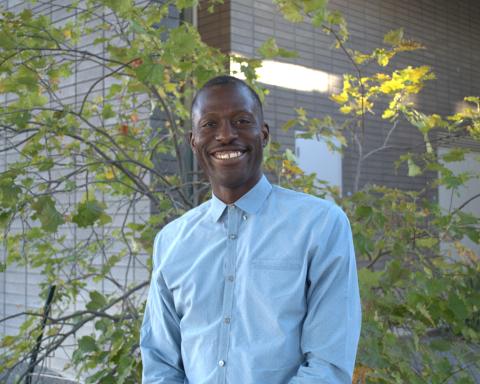 Roger Reeves wears a light blue button-down shirt and stands in front of a mid-sized leafy green tree. A grey brick building is also visible in the background.