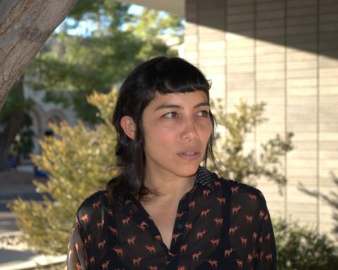 Tilsa Otta wears a black button-down shirt printed with small deer and a polka-dot collar. She stands before a grey brick building and a large green bush.