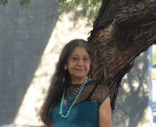 Lorna Dee Cervantes poses in front of a mesquite tree in a shaft of sunlight. She wears a teal top with black mesh, dangly earrings, and two necklaces: one that is blue, white, and beaded, and another with small turquoise stones.