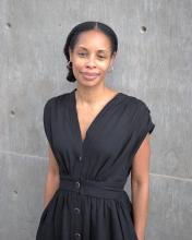 Nicole Sealey stands in front of a gray wall and wears a black dress. 