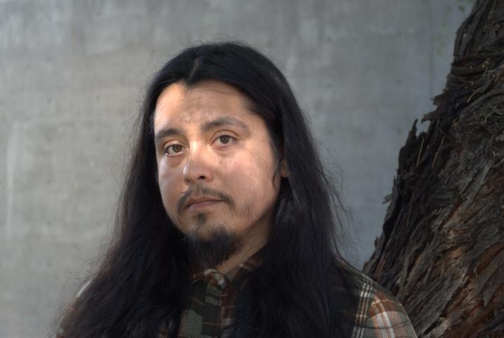 Angel Dominguez, a poet with long dark hair and a mustache, stands next to a mesquite tree in golden hour light, some of which lands on their face.