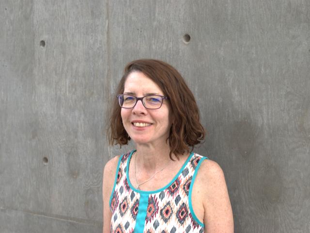 Clare Sullivan wears a multicolored sleeveless blouse and dark purple wire-framed classes. She stands against a gray wall.