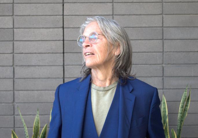 Eileen Myles looks off to the side, not directly at the camera, and wears a navy blue blazer and vest with an olive green shirt. They are standing in front of a grey wall, and there are some plants visible in the background.
