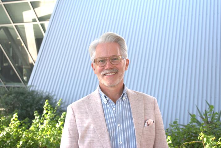 Mark Wunderlich wears a tan blazer over a pale blue button-down with vertical white stripes, as well as glasses with tan, translucent frames. He stands in front of a white, sloped roof with ridges that is next to a glass wall, also sloped. There are also shrubs visible in the background of the photo.