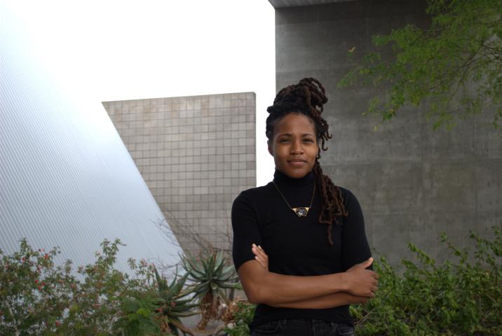 Saretta Morgan stands in a black turtleneck t shirt and grey gemstone necklace with arms slightly crossed. She stands outside in front of grey walls, a slanted white wall, and shrubs.