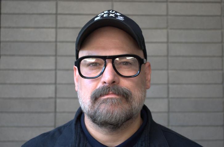 Timothy Donnelly wears a black baseball cap, black-rimmed glasses, and stands before a gray-wall background.