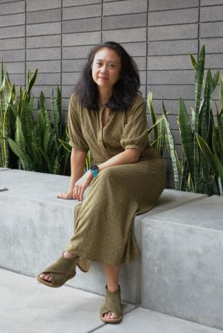 Jennifer Chang sits on a grey stone slab bench. She wears a mustard yellow quarter-sleeve length jumpsuit with a black geometric pattern, and matching suede mustard yellow sandals. She sits in front of a grey brick wall with a row of aloe vera plants behind her.