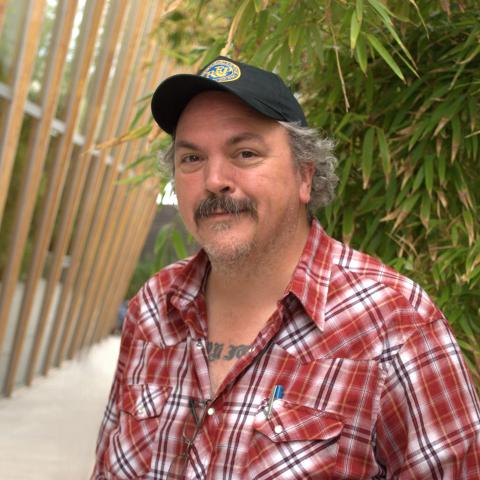 Mathias Svalina wears a red and white plaid shirt and a black baseball cap. He stands in front of a grove of bamboo trees and a wood-paneled glass wall.