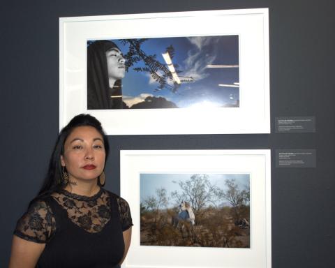 Su:k Chu:vak Fulwider wears red lipstick, a floral, black lace shirt, and circular earrings. She stands next to her photographs framed on a dark gray wall, one of which features a desert landscape with a white horse, and another with a boy with closed eyes against a blue sky and mesquite leaves. 