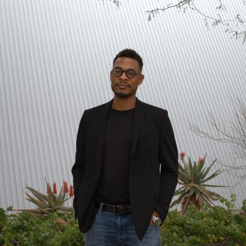 Terrance Hayes wears a black sweater and a black blazer with blue jeans and a black belt. He also has on black-rimmed glasses. Hayes stands in front of a white background, with aloe plants in the background flaunting orange blooms.