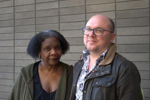 Vievee Francis wears a dark green sweater, black dress, and gold earrings. Matthew Olzmann wears a navy and white paisley shirt, dark brown jacket, and navy blue glasses. They pose standing next to each other in front of a grey brick wall. 