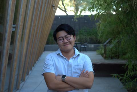 Yanyi stands, smiling, with arms crossed in the Poetry Center's back garden that consists of bamboo and a rock slab. The Poetry Center's slightly curved back wall made of glass and wooden slabs frames the left side of the photo.