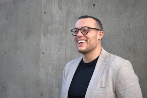 Poet Charif Shanahan stands in front of a slate gray wall wearing dark-rimmed glasses, a grey blazer, and a black t-shirt. He laughs as he faces the camera at a slight angle.