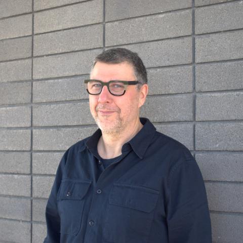 Matthew Zapruder wears a navy button-down shirt and glasses with olive green frames. He stands before a grey brick wall.