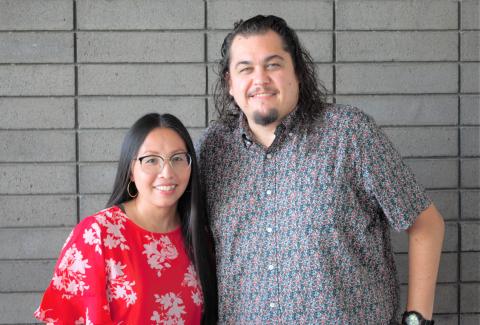 Mai Der Vang, wearing a red and white dress, and Anthony Cody, wearing a pattered button-down, stand together in front of a grey brick wall.