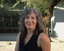 Susan Briante wears a black sleeveless shirt and gold necklaces. She poses in front of a mesquite tree and a glass wall of a building.