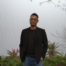 Terrance Hayes wears a black sweater and a black blazer with blue jeans and a black belt. He also has on black-rimmed glasses. Hayes stands in front of a white background, with aloe plants in the background flaunting orange blooms.