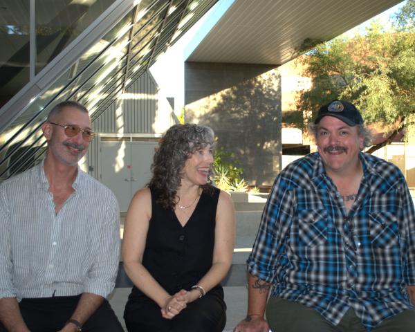 Farid Matuk, Susan Briante, and Mathias Svalina sit together in a row. Farid wears glasses and a grey and white striped button down and black pants. Susan wears a black sleeveless top and black pants. Mathias wears a baseball cap and a blue and grey plaid shirt with grey pants. They sit together in front of a glass-paneled wall and a grey stone patio area.