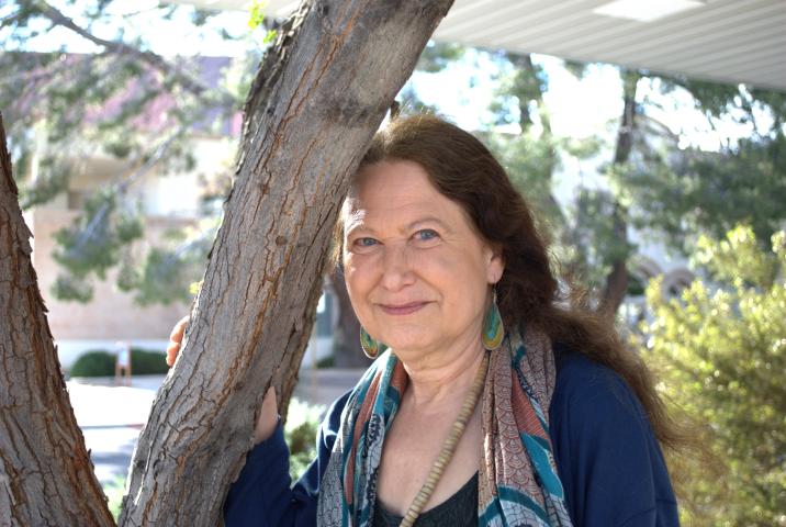 Jane Hirshfield leans against a tree and wears a blue top with a multicolored scarf, as well as earrings and a beaded necklace.
