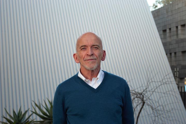Mark Doty wears a blue sweater over a white shirt and stands in front of a white wall with plants visible behind him.