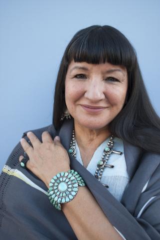 Sandra Cisneros wears a grey shawl and turquoise bracelet while smiling in front of a light blue background