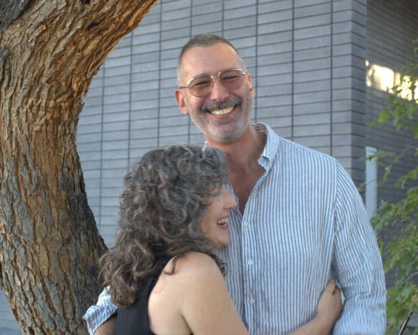 Susan Briante and Farid Matuk stand in embrace beneath a mesquite tree. Susan wears a black sleeveless top, and Farid wears glasses and a gray and white striped button-down. They stand in front of a grey brick wall.