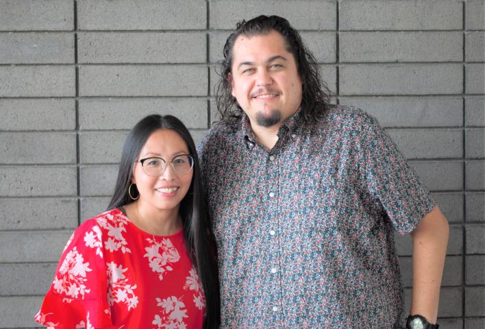 Mai Der Vang, wearing a red and white dress, and Anthony Cody, wearing a pattered button-down, stand together in front of a grey brick wall.